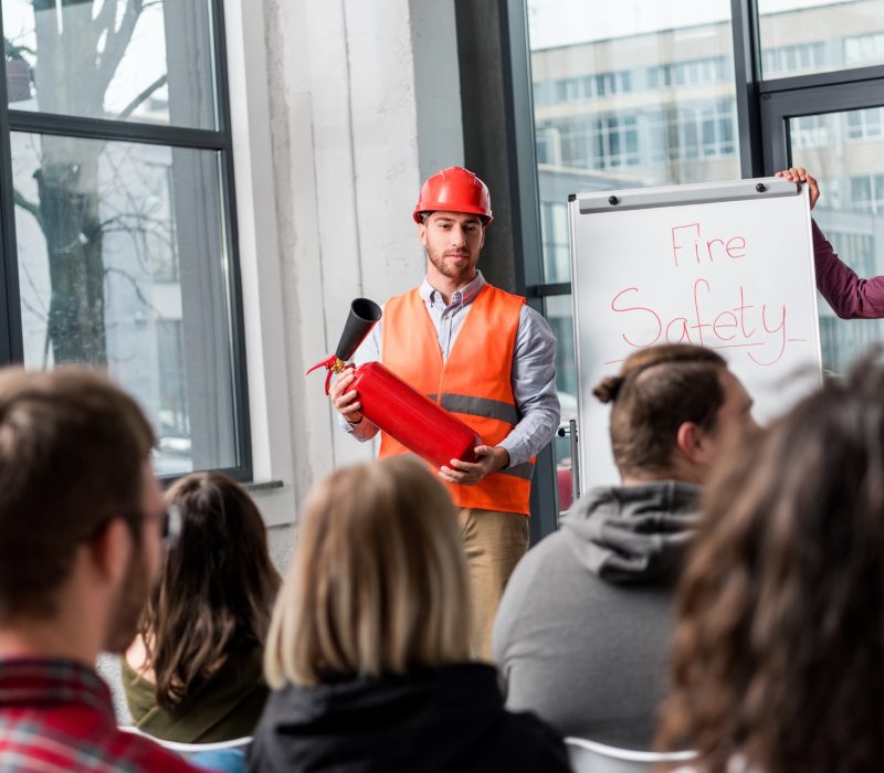 selective focus of handsome fireman in helmet holding extinguisher while standing near white board